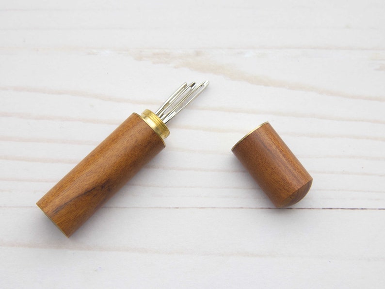 May include: A cylindrical wooden needle case with a brass screw-top lid, shown open to reveal several silver-toned metal sewing needles stored inside. The case features a smooth, polished brown wood grain finish and a metallic brass threaded neck.