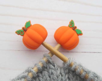 May include: Two orange pumpkin-shaped stitch markers with green leaves and stems are crossed over a gray knitted project. The markers are on wooden knitting needles, perfect for fall-themed crafts.