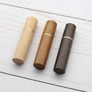 May include: Three cylindrical wooden cases arranged in a row, each featuring a distinct wood tone ranging from light to dark brown. Each case is divided into two sections by a thin, horizontal gold-colored metal band encircling the middle.