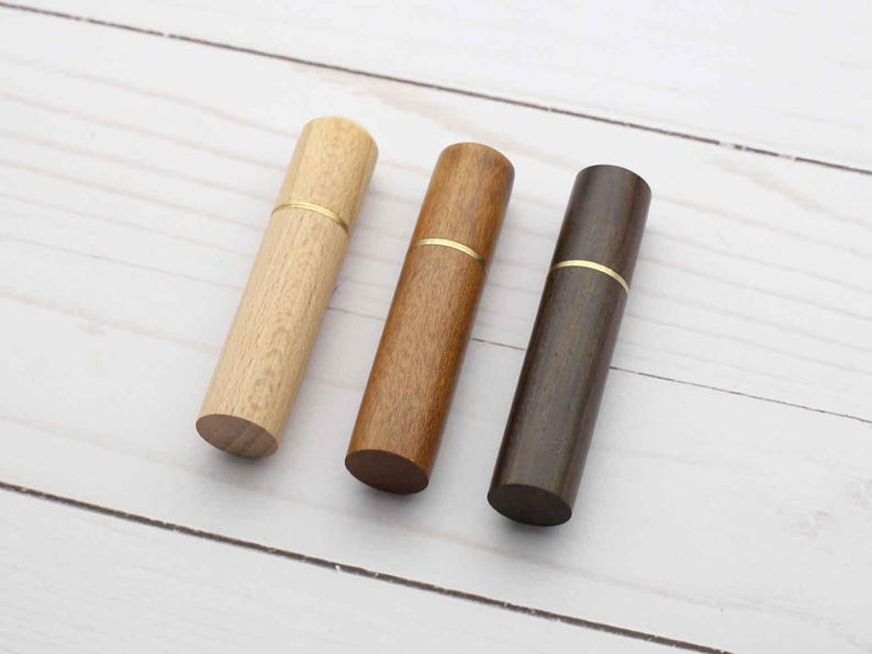 May include: Three cylindrical wooden cases arranged in a row, each featuring a distinct wood tone ranging from light to dark brown. Each case is divided into two sections by a thin, horizontal gold-colored metal band encircling the middle.
