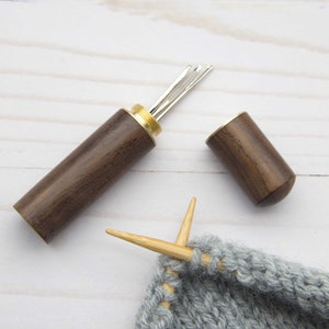 May include: A dark brown wooden needle case with a brass screw-top lid, containing several silver metal sewing needles. The case is cylindrical with a smooth finish. Beside it, two light-colored wooden knitting needles are inserted into grey yarn.