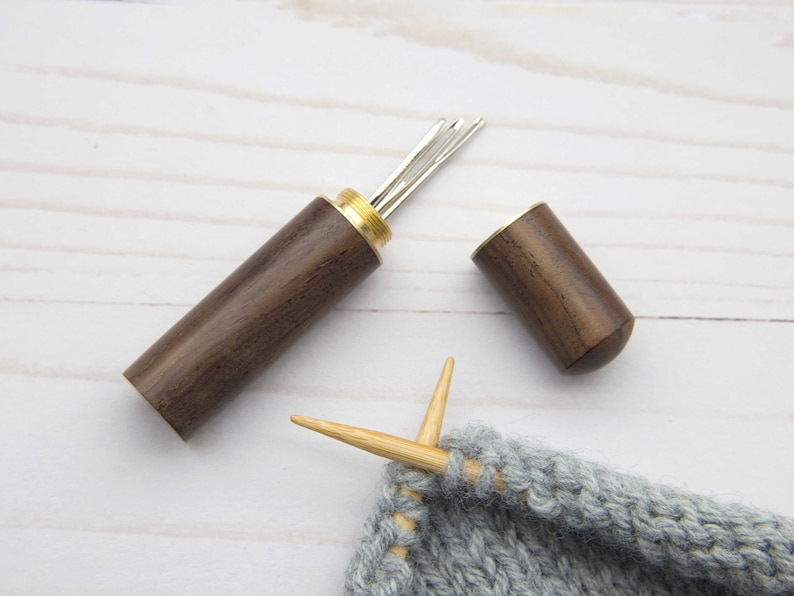 May include: A dark brown wooden needle case with a brass screw-top lid, containing several silver metal sewing needles. The case is cylindrical with a smooth finish. Beside it, two light-colored wooden knitting needles are inserted into grey yarn.
