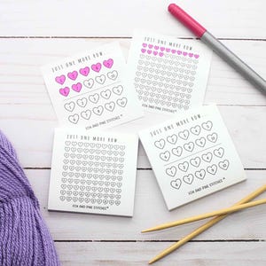 May include: Three square white paper sticky note pads with the text JUST ONE MORE ROW and FOX AND PINE STITCHES. Each pad features a grid of numbered heart outlines. One pad has seven hearts filled with pink ink. A pink marker and two wooden knitting needles.