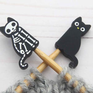 May include: Two black cat-shaped stitch markers with bamboo needles crossed over grey yarn. One cat is a skeleton design, the other is a solid black cat with yellow eyes. A fun accessory for knitting projects.