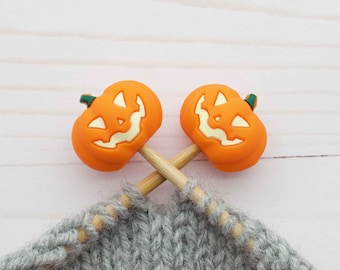 May include: Two wooden knitting needles with orange pumpkin-shaped ends, each with a carved jack-o'-lantern face. The needles are crossed over a gray knitted fabric.