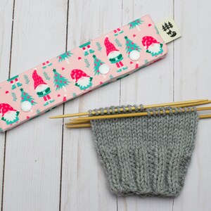 May include: A pink knitting needle case with a Christmas gnome and tree pattern, secured with white snaps. Beside it, a gray knitted sock is in progress, held by wooden knitting needles. The scene is set on a light wood surface.