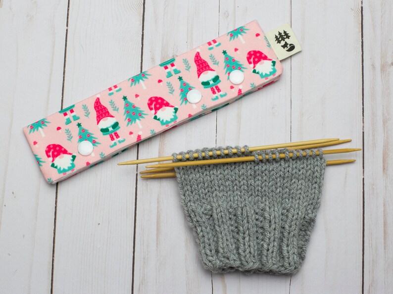 May include: A pink knitting needle case with a Christmas gnome and tree pattern, secured with white snaps. Beside it, a gray knitted sock is in progress, held by wooden knitting needles. The scene is set on a light wood surface.