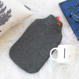 Może przedstawiać: Szara butelka na gorącą wodę z dzianinową szyją, biały kubek z literą "L", niebieski notatnik i dekoracyjna poduszka z motywem drzewa. Przedmioty ułożone są na białej, puszystej powierzchni.