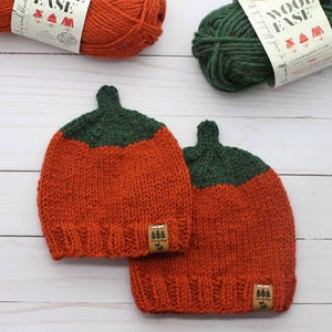 May include: Two hand-knitted beanies featuring a pumpkin-inspired design with a vibrant orange body and a dark green ribbed top section. Each hat includes a small brown rectangular tag with a tree icon and the text Pine & Pine stitched on the brim.