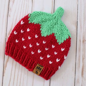 Puede incluir: Un gorro de fresa tejido en rojo y verde, con detalles de semillas blancas. El gorro tiene un tallo verde y una peque&ntilde;a etiqueta marr&oacute;n. El gorro est&aacute; hecho de hilo suave y est&aacute; dise&ntilde;ado para parecerse a una fresa.