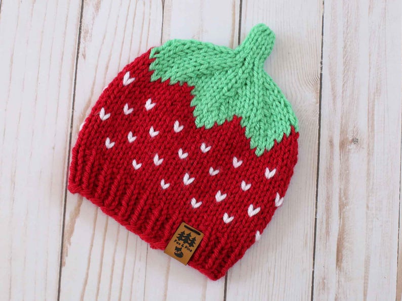 Puede incluir: Un gorro de fresa tejido en rojo y verde, con detalles de semillas blancas. El gorro tiene un tallo verde y una peque&ntilde;a etiqueta marr&oacute;n. El gorro est&aacute; hecho de hilo suave y est&aacute; dise&ntilde;ado para parecerse a una fresa.