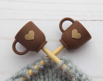May include: Two brown coffee cup-shaped stitch markers with tan heart accents are crossed over knitting needles. The needles are inserted into a gray knitted fabric, suggesting a knitting project in progress. The background is a light wood surface.