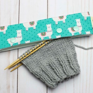May include: A teal blue fabric headband with a white llama print and white polka dots. The headband has two light blue buttons. The headband is lying on a white wooden surface with a gray knitted fabric and two wooden knitting needles.