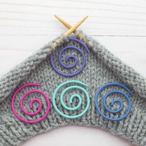 May include: Four colorful spiral stitch markers on a gray knitted fabric with wooden knitting needles.