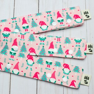 May include: Three fabric wristbands with a pink background and a repeating pattern of gnomes and Christmas trees in red and teal. Each band has a white snap closure and a small tag with a forest and fox logo.