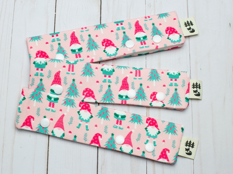 May include: Three fabric wristbands with a pink background and a repeating pattern of gnomes and Christmas trees in red and teal. Each band has a white snap closure and a small tag with a forest and fox logo.