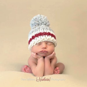 May include: A newborn baby wearing a gray and white knitted hat with a red stripe and a pom-pom on top. The baby is sleeping with their hands clasped together under their chin.
