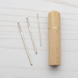 May include: Three silver-colored metal sewing needles with large eyes arranged next to a cylindrical light-brown wooden needle case. The case features a smooth grain texture and a thin gold-colored metal band separating the lid from the base.