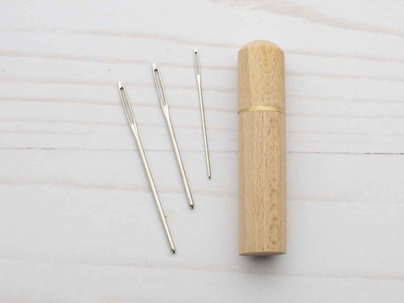 May include: Three silver-colored metal sewing needles with large eyes arranged next to a cylindrical light-brown wooden needle case. The case features a smooth grain texture and a thin gold-colored metal band separating the lid from the base.