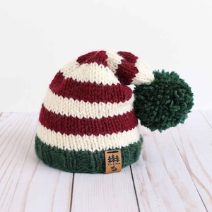 May include: A knitted hat with burgundy and white stripes, finished with a green pom-pom. The hat has a green brim and a small brown tag with a logo. The hat is designed for cold weather.