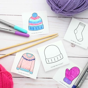 May include: Square cards featuring line art of a beanie, sock, sweater, and mitten, with some colored in. The text "FOX AND PINE STITCHES" is printed on each. Two wooden knitting needles, two balls of yarn in purple and pink, and markers are present.