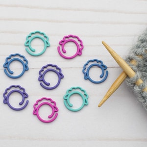 May include: A set of eight colourful stitch markers in the shape of a sheep. The markers are in shades of blue, purple and pink. A pair of knitting needles and a grey knitted fabric are also in the image.