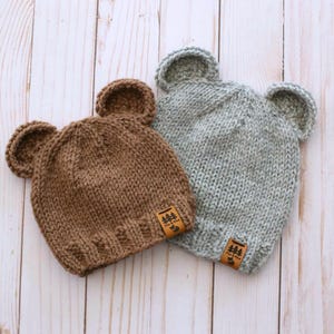 Peut inclure: Deux bonnets en tricot en forme d'ours. L'un est marron, l'autre gris. Chaque bonnet a deux oreilles arrondies et une petite étiquette marron. Les bonnets sont en tricot épais et sont conçus pour les bébés ou les enfants.