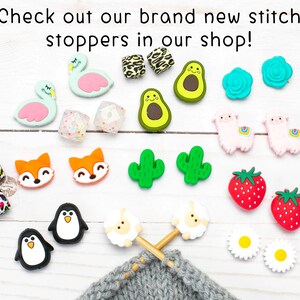 May include: A selection of colourful silicone stitch markers, featuring flamingos, avocados, cacti, strawberries and sheep. The markers are shown on a white wooden surface with a knitting project in the background.