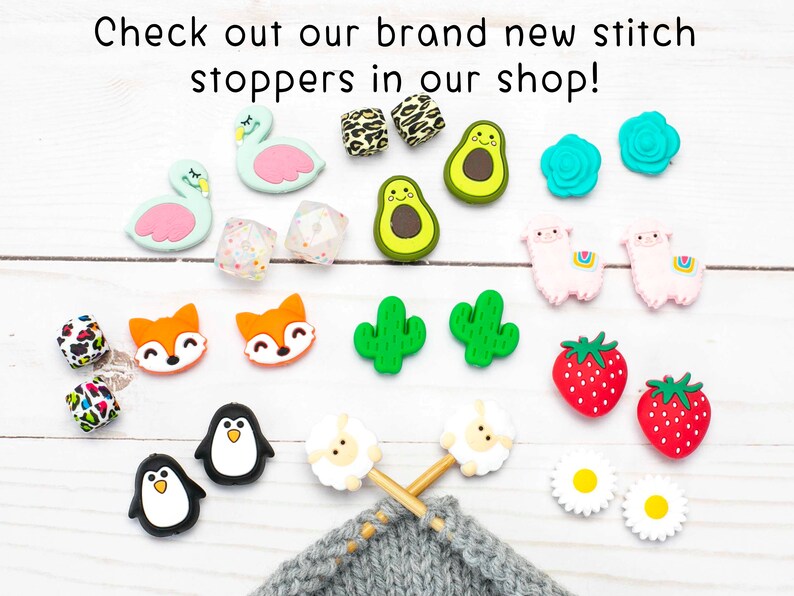 May include: A selection of colourful silicone stitch markers, featuring flamingos, avocados, cacti, strawberries and sheep. The markers are shown on a white wooden surface with a knitting project in the background.
