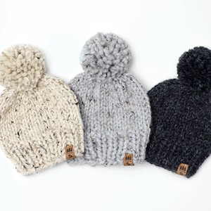 May include: Three knitted beanies with pom-poms in beige, grey, and black. Each beanie has a small brown leather patch with a tree logo.