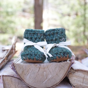 May include: A pair of handmade, dark green crochet baby booties with white ribbon ties. The booties have brown leather soles and are displayed on a stack of wood logs, suggesting a rustic, natural theme.