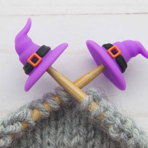 Könnte beinhalten: Zwei Stricknadeln mit einem grauen Strickprojekt sind mit lila Hexenhut-Maschenmarkierern versehen. Die Hüte haben schwarze Bänder mit orangefarbenen Schnallen. Der Hintergrund ist eine weiße Holzoberfläche.