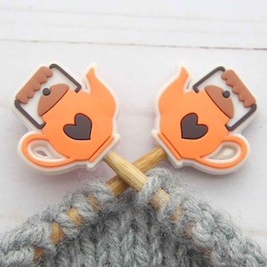 Op de afbeelding: Twee oranje theepotvormige steekmarkeerders met een hartontwerp, op houten breinaalden. De markeerders staan tegen een achtergrond van grijze gebreide stof, ideaal voor handwerk.