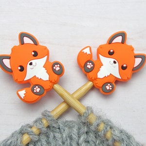 May include: Two orange fox-shaped knitting needle stoppers on a pair of wooden knitting needles. The needles are resting on a gray knitted fabric.