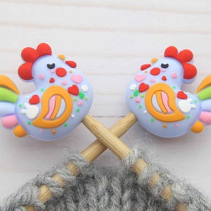 Può includere: Due segnapunti a forma di gallina blu chiaro con creste rosse e accenti colorati sono incrociati su un progetto a maglia grigio. I segnapunti hanno dettagli arancioni, rosa e verdi. Lo sfondo è una superficie di legno chiaro.