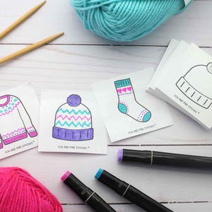 Puede incluir: Un conjunto de tarjetas cuadradas con ilustraciones de un suéter, un gorro, un calcetín y un contorno de gorro en blanco, cada una etiquetada como Fox and Pine Stitches. Las tarjetas se muestran con dos agujas de tejer de madera, dos ovillos de lana y marcadores.