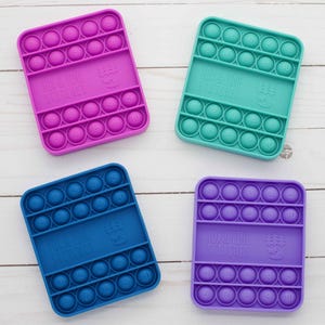 May include: Four square silicone fidget toys in different colors: pink, blue, teal, and purple. Each toy has a grid of circles that can be pushed in and out, and the text "PUSH & POP FIDGET TOYS" is printed on each toy.