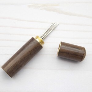 May include: A cylindrical dark brown wooden needle case with a brass screw-top lid. The case is open, revealing several silver-colored metal sewing needles stored inside the brass-threaded opening of the main wooden body.