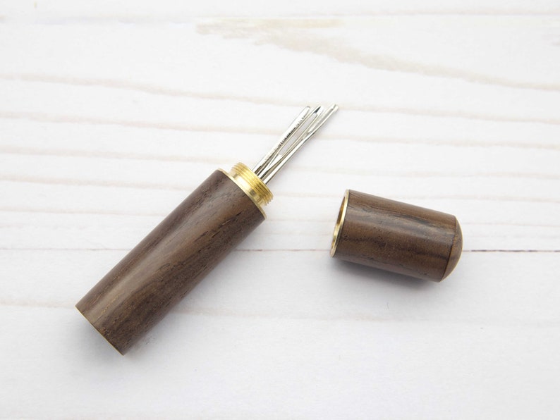 May include: A cylindrical dark brown wooden needle case with a brass screw-top lid. The case is open, revealing several silver-colored metal sewing needles stored inside the brass-threaded opening of the main wooden body.