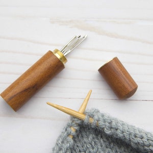 May include: A cylindrical wooden needle case with a brass screw top, shown open with several silver metal needles inside. Beside the case lies a matching wooden cap and a piece of grey knitted fabric with two light-colored knitting needles.