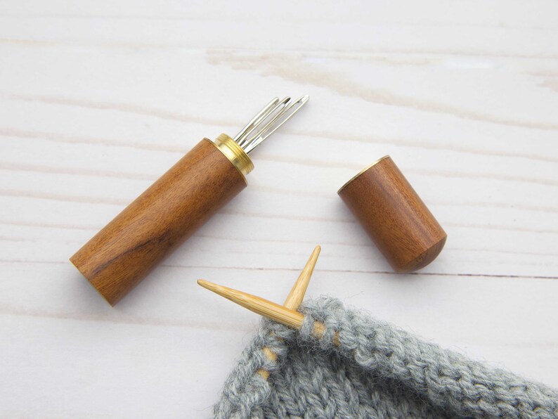 May include: A cylindrical wooden needle case with a brass screw top, shown open with several silver metal needles inside. Beside the case lies a matching wooden cap and a piece of grey knitted fabric with two light-colored knitting needles.