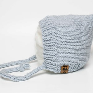 Könnte beinhalten: Eine hellblaue gestrickte Babyhaube mit langem Band. Die Haube hat ein kleines braunes Leder-Etikett mit dem Text "Pine & Pine" und einer kleinen Baumgrafik.