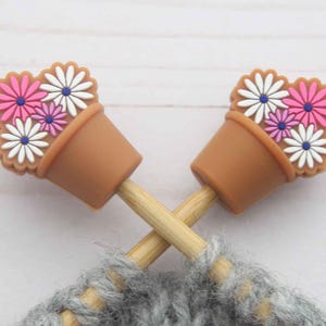 Può includere: Due puntali per ferri da maglia con vasi di fiori marroni e fiori colorati. I fiori sono bianchi, rosa e viola. Gli aghi sono incrociati su un pezzo di maglia grigio.