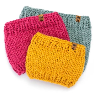 May include: Three chunky knit headbands in shades of fuchsia, mustard yellow, and teal. Each headband has a ribbed texture and a small brown tag with a tree logo. The headbands are displayed on a white background.