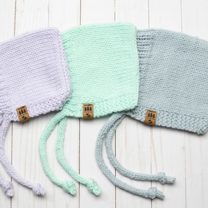 Può includere: Tre cappellini di lana per neonati con lacci in lavanda, verde menta e grigio chiaro. Ogni cappellino ha un'etichetta in pelle marrone con il testo "Pink & Blue" e un piccolo grafico di un albero.