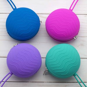 Peut inclure: Quatre pochettes rondes à fermeture éclair de différentes couleurs : bleu, rose, violet et turquoise. Chaque pochette présente un motif ondulé et une petite boucle pour l’accrocher.