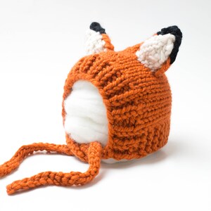 Op de afbeelding: Een gebreide oranje vossenmuts met witte en zwarte accenten. De muts heeft een lange oranje strik.