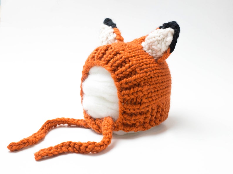 Op de afbeelding: Een gebreide oranje vossenmuts met witte en zwarte accenten. De muts heeft een lange oranje strik.