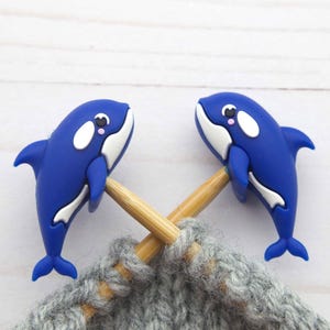 May include: Two blue orca-shaped stitch markers with white accents and pink cheeks are attached to wooden knitting needles, resting on gray knitted fabric. The orcas are facing each other. The background is a white wooden surface.