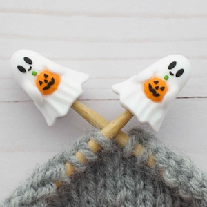 May include: Two white ghost-shaped stitch markers with orange pumpkin details are crossed over a gray knitted fabric. The markers are on wooden knitting needles. The background is a light wood surface. Halloween-themed knitting accessories.